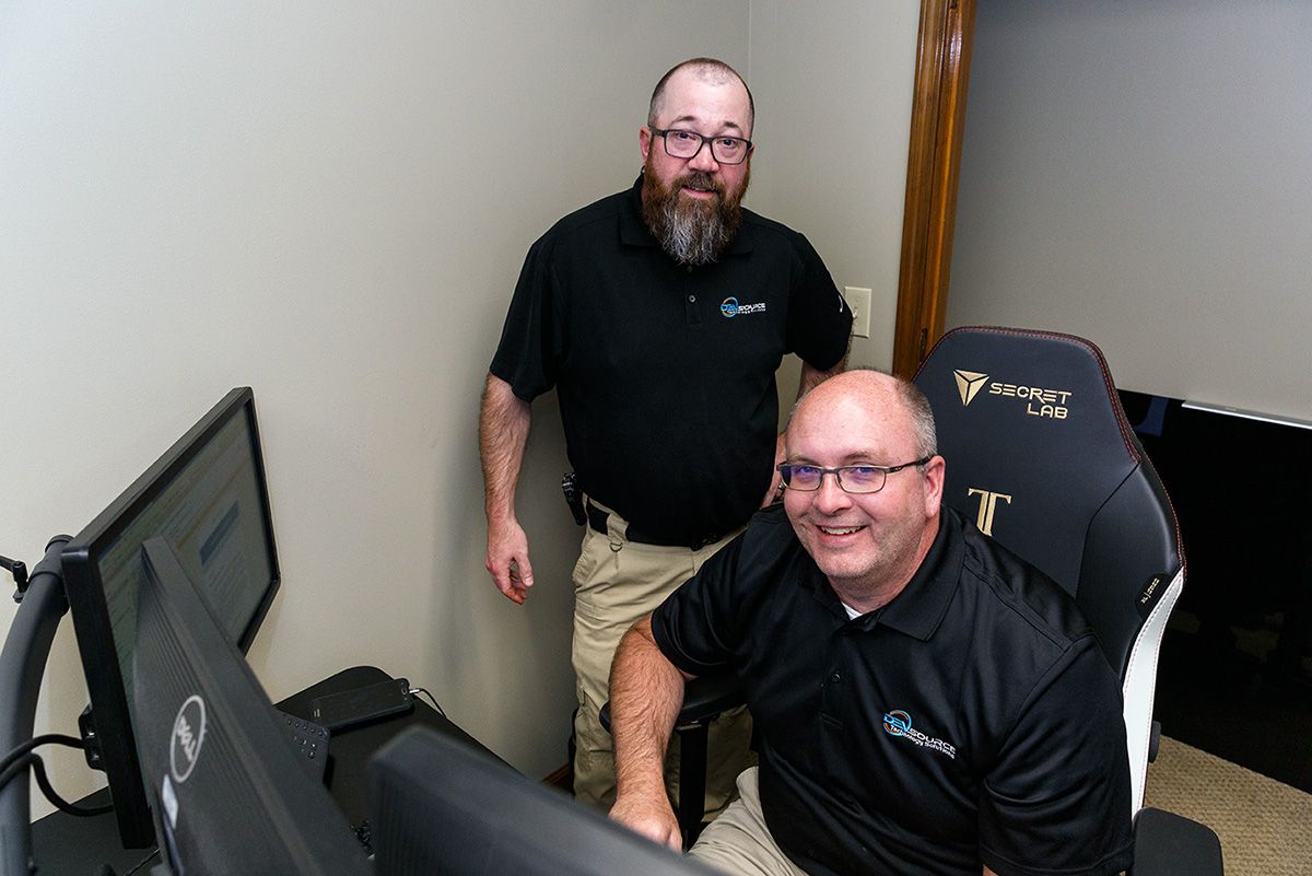 Two DEVsource employees, one sitting and one standing, smiling at the camera